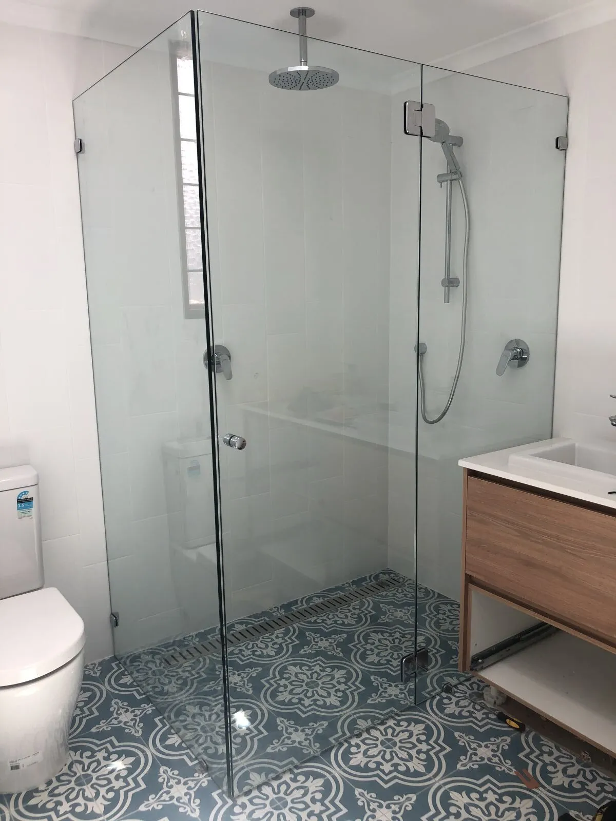 Frameless shower screen beside a timber vanity on patterned tiles